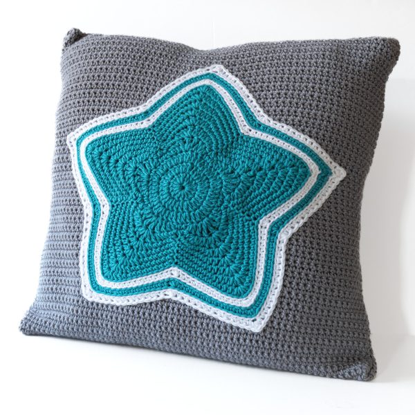 a crochet cushion worked in dark grey featuring a teal and white star at the centre on a plain white background. winter star cushion crochet pattern